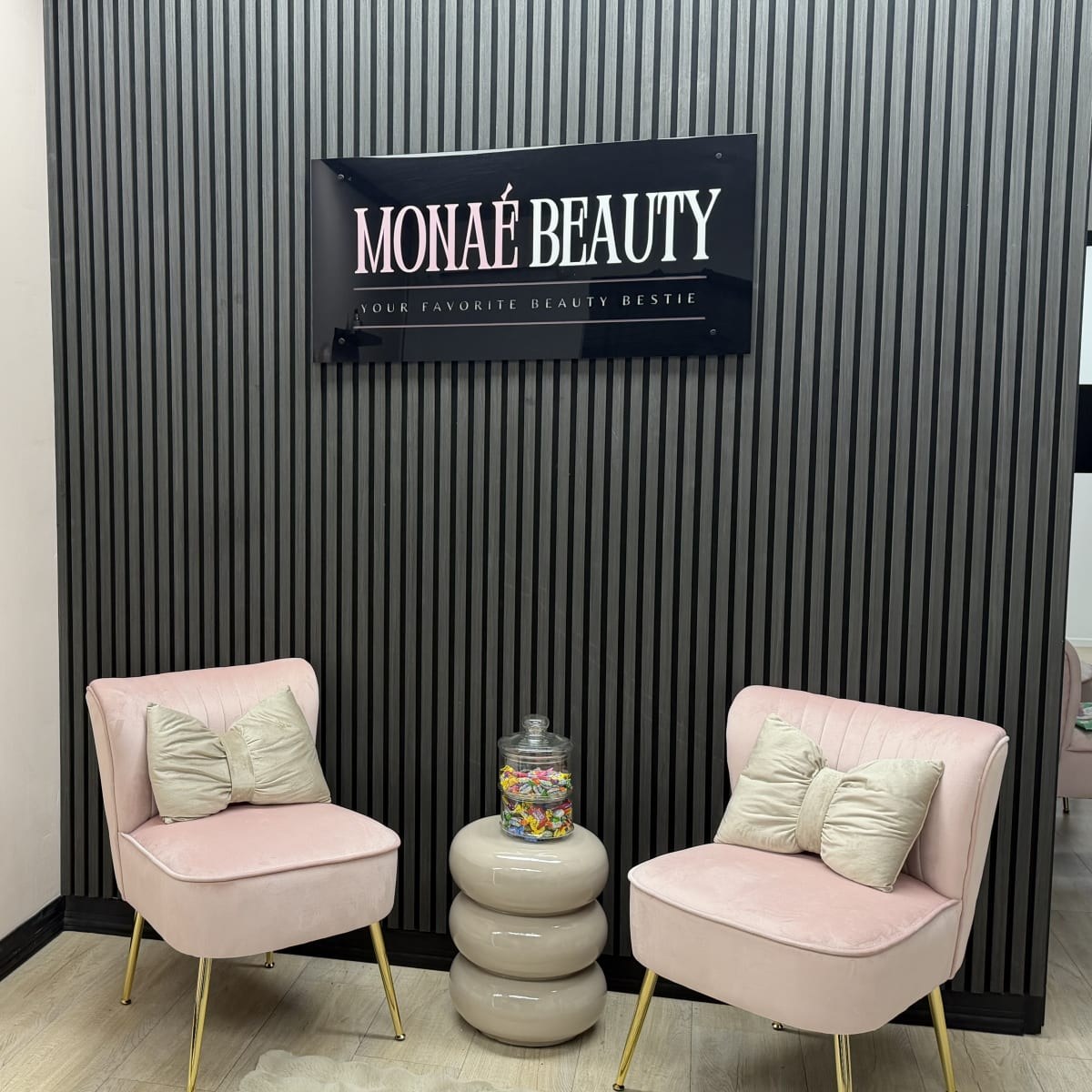 De wachtruimte van Monaé Beauty: een zwarte lamellenwand met het logo, twee roze fluwelen stoeltjes en een snoeppot